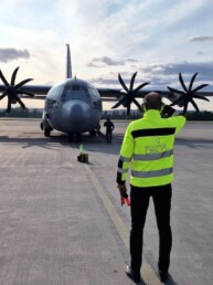 Man in yellow jacket is marshalling a four propelled gray military plane at Gardermoen Airport.