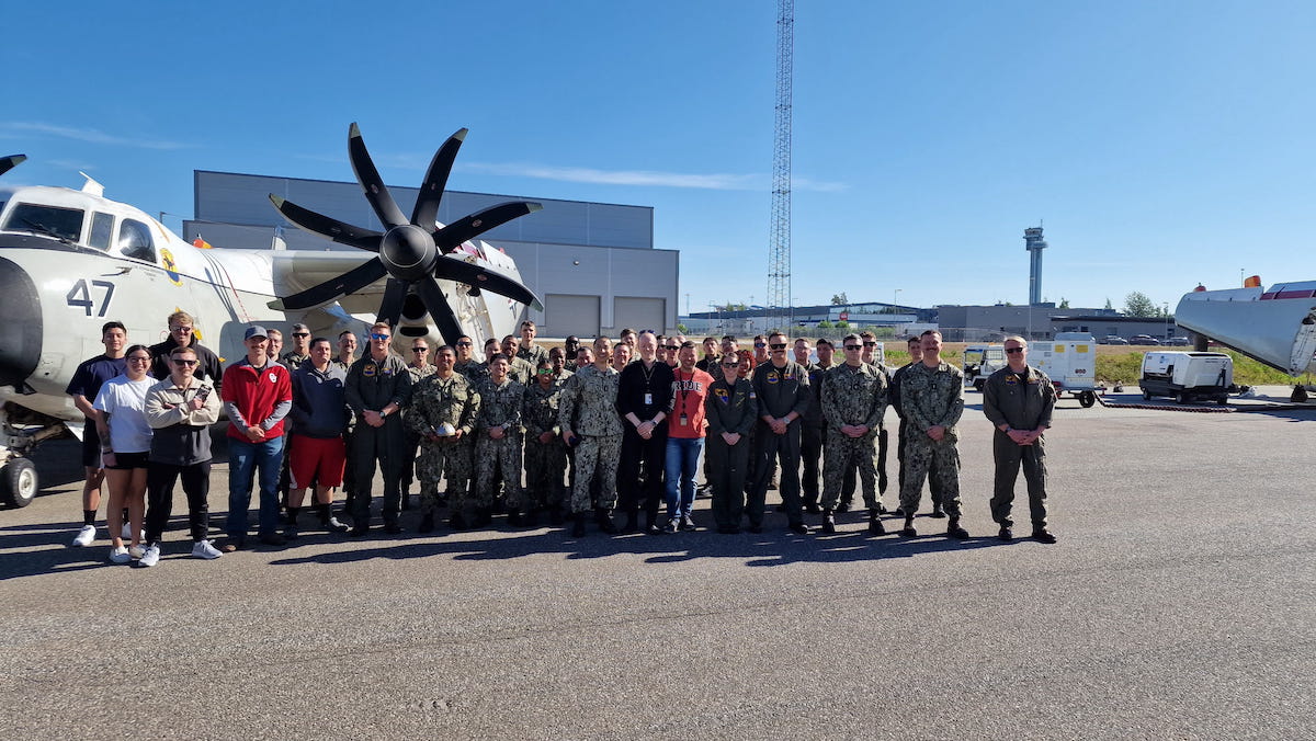 A big group of people, half of them in military uniforms, is standing in front of a military pane. They're smiling, it's a sunny day.