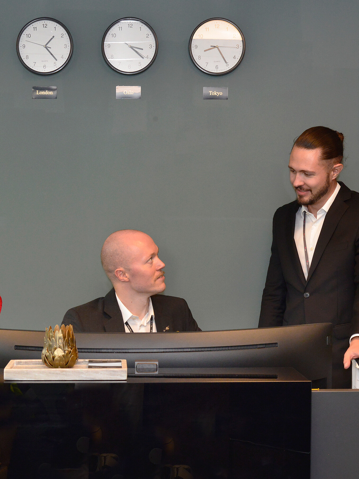 Members of staff at our FBO in Torp is positioned behind the reception desk, looking at each other. Clocks on the wall show Oslo, London and Tokyo.